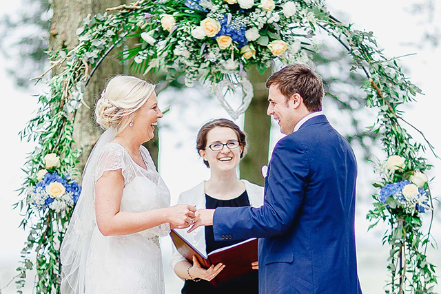 A bride and groom hold hands underneath a flower arch, in front of Stephanie